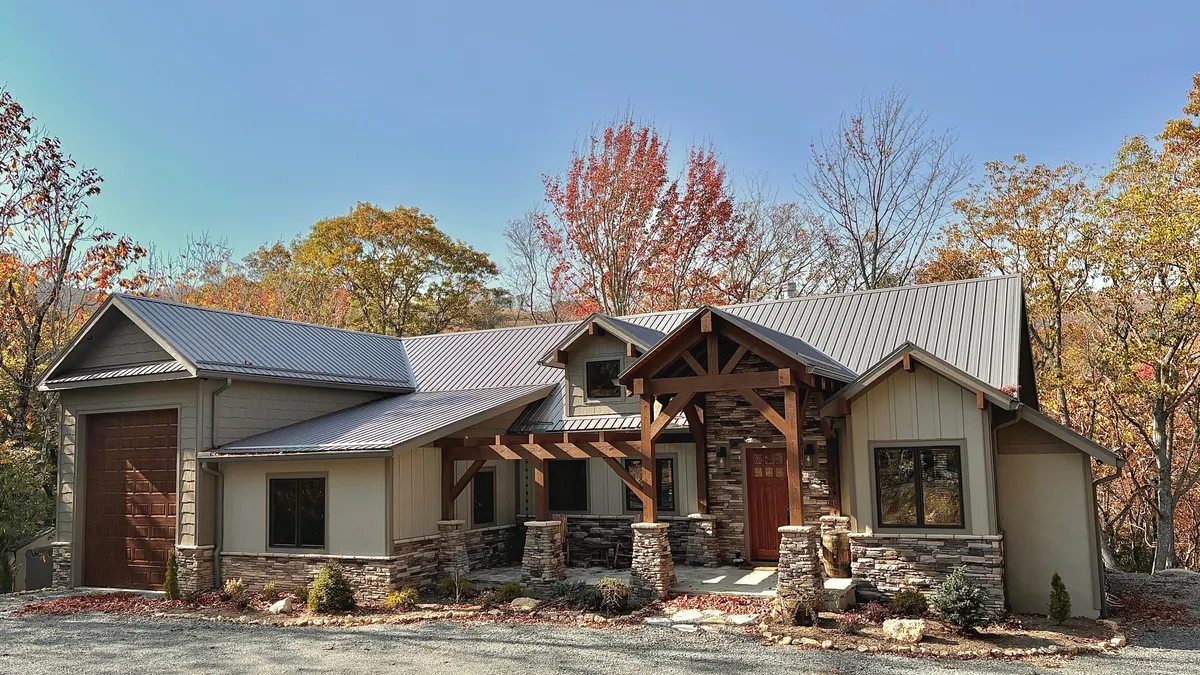 The image showcases the grand wooden entrance and stone façade of a modern single-family cabin, set against vibrant autumn foliage. The spacious front porch invites guests to relax and enjoy the tranquil mountain setting, hinting at the luxury and coziness found inside.