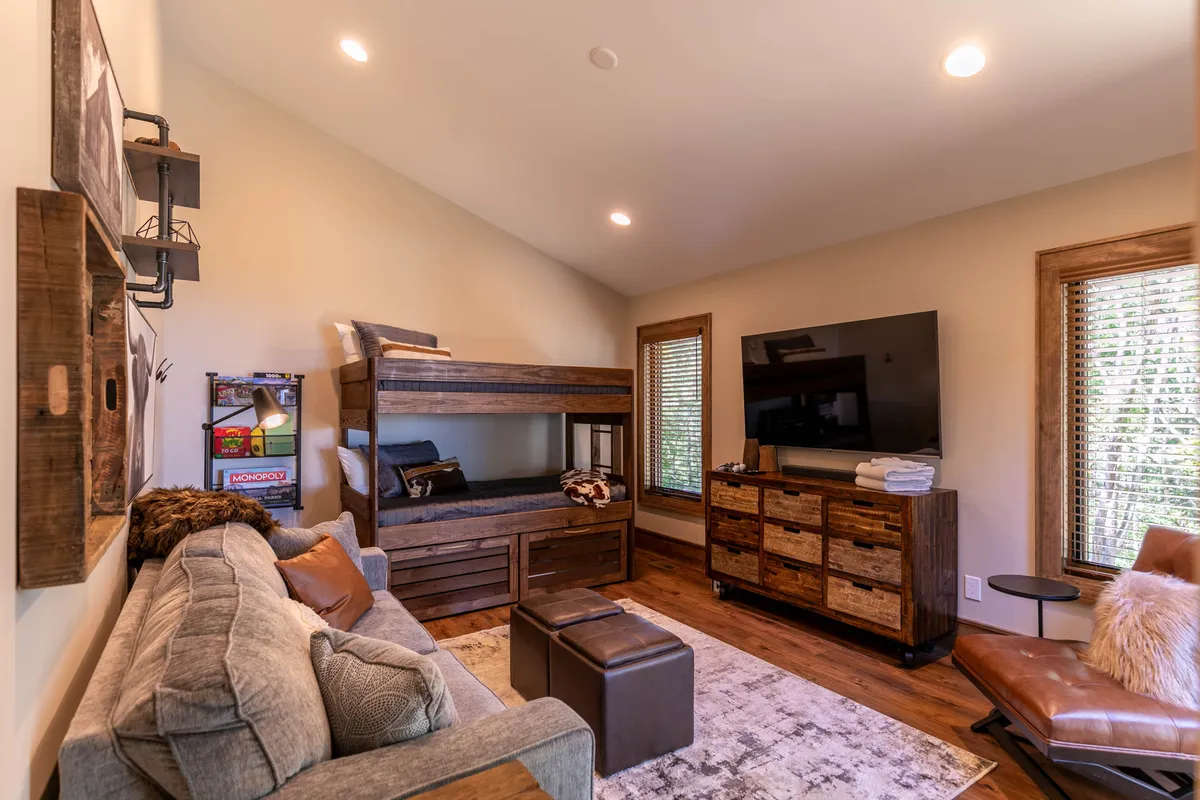 A playful yet functional bedroom/den outfitted with a twin-over-twin bunk bed featuring built-in storage, a comfortable sofa that doubles as a queen sleeper, a TV, and wooden furniture. The mood is inviting and versatile, perfect for families or groups seeking a multi-purpose sleeping space.