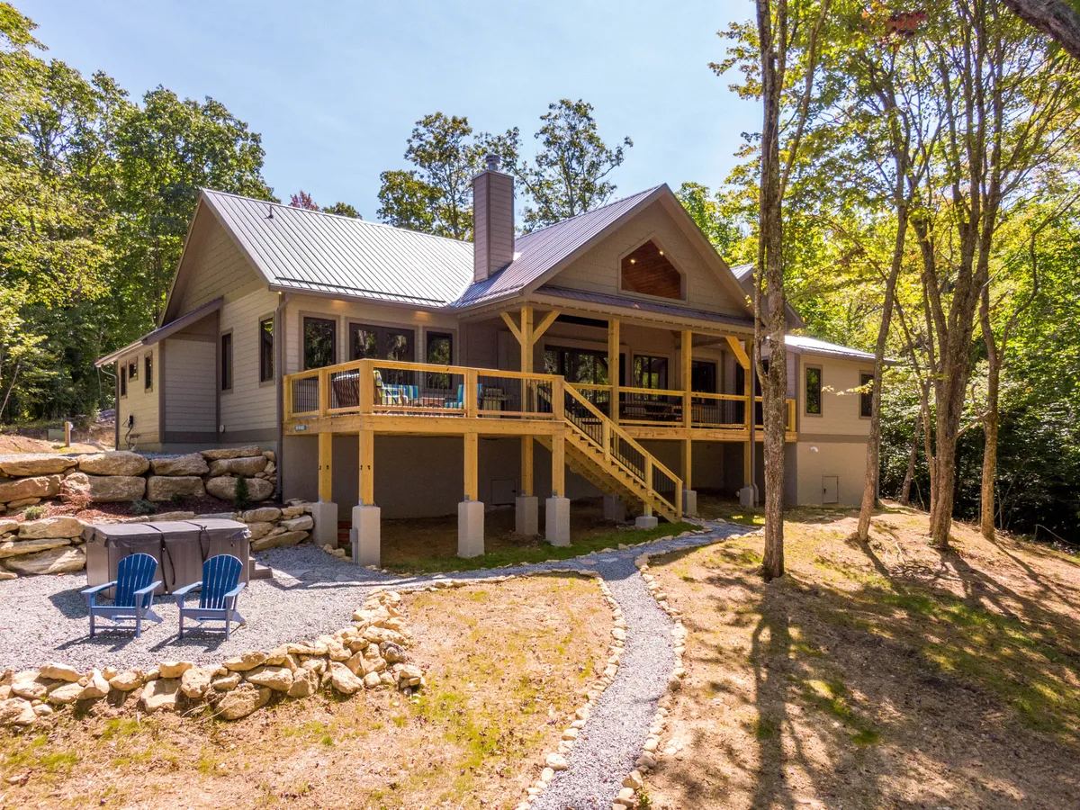 A wide view of the spacious wooden deck featuring a stone fire pit surrounded by six blue Adirondack chairs, natural stone landscaping, and lush greenery in the background. The deck offers plenty of space for both dining and lounging, blending seamlessly with the mountain setting.