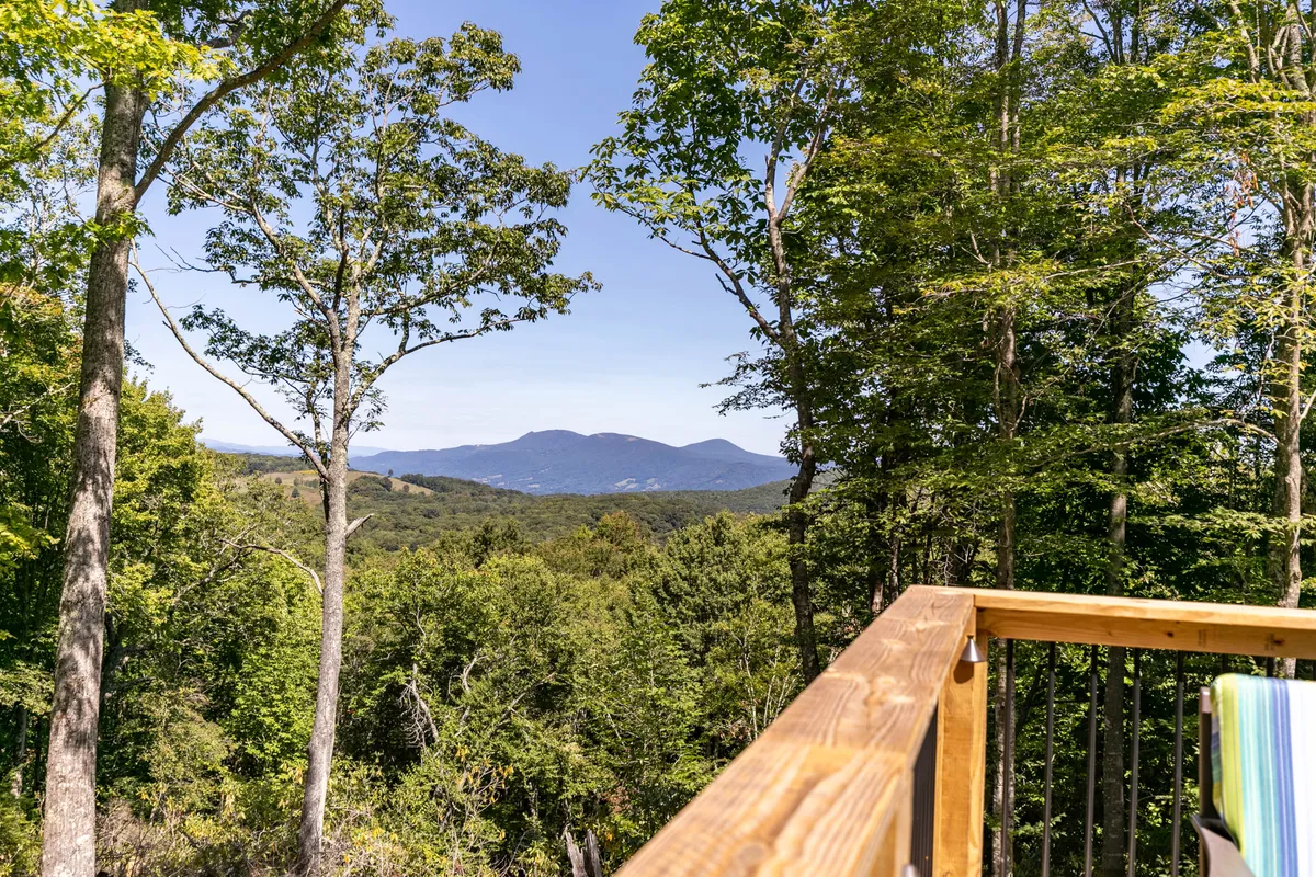 A breathtaking view from the deck, overlooking a distant mountain range surrounded by lush green trees and framed by a wooden railing. The scene is serene and invites guests to immerse themselves in peaceful mountain scenery.