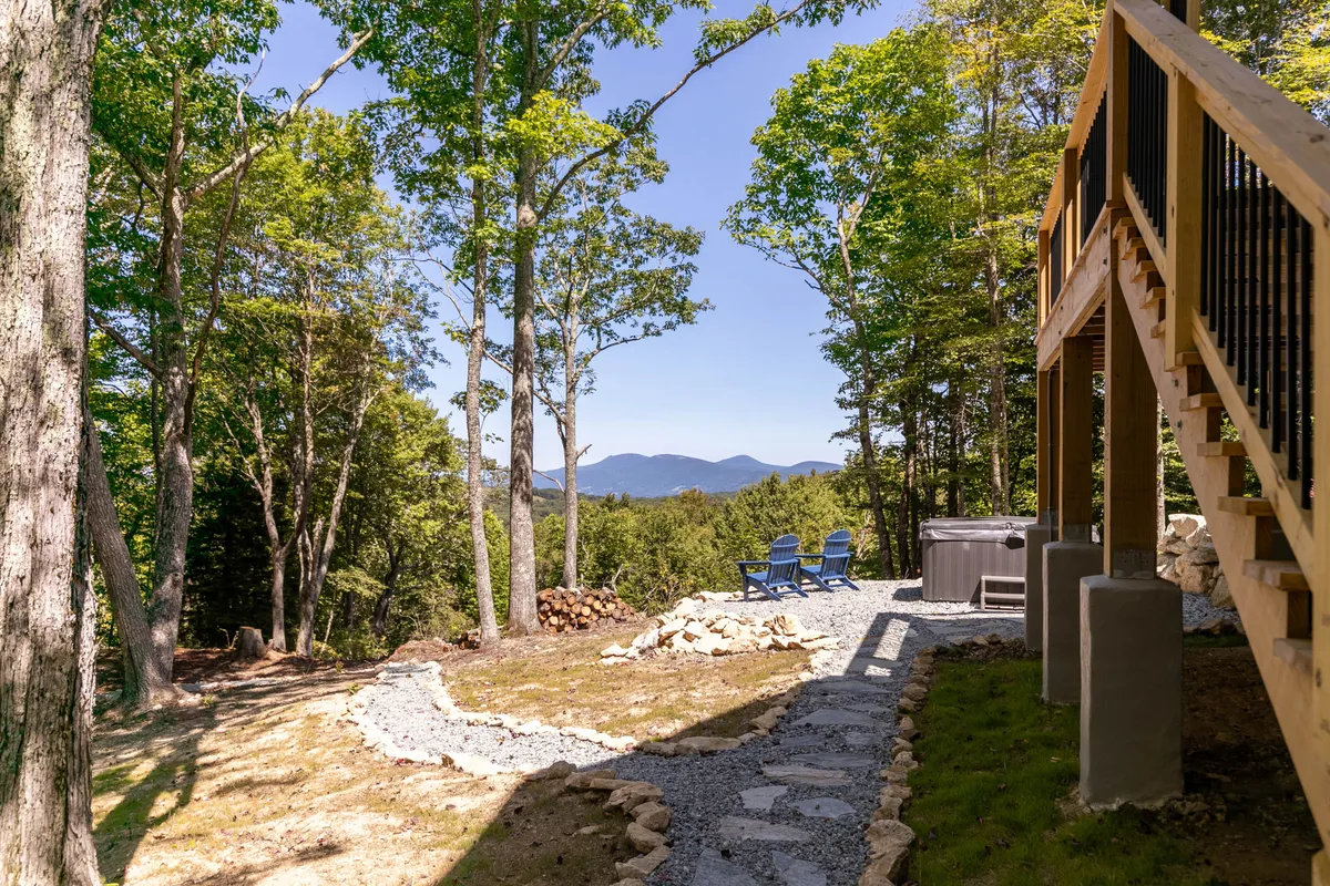 This image features a scenic hot tub area with blue chairs, a clear view of the mountains, and a pathway leading through a tranquil natural landscape. The hot tub is positioned for optimal enjoyment of sunrise, sunset, and stargazing moments, surrounded by the peaceful beauty of the mountain setting.