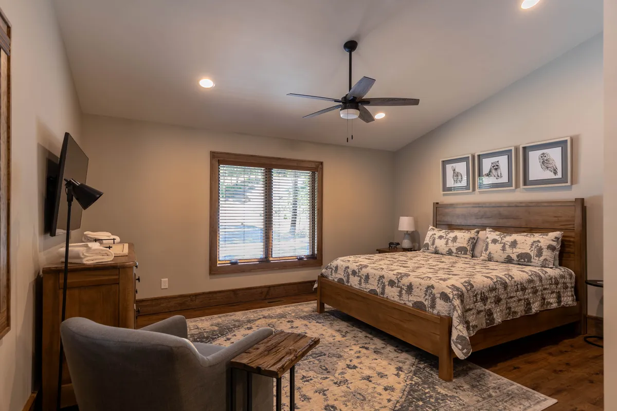 A tranquil, cozy king suite featuring a bed with patterned bedding, wooden furniture, a window with blinds allowing in soft natural light, an armchair perfect for reading, and tasteful artwork on the wall. The composition radiates calm and comfort, ideal for restful sleep and private downtime.