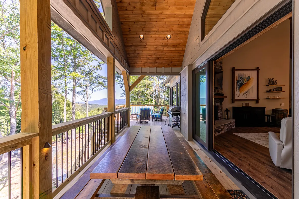 A wooden outdoor dining table and seating area on a deck, with sweeping mountain views and easy access to the indoors through sliding glass doors. The scene suggests relaxed al fresco dining and communal gatherings, with nature as the backdrop.