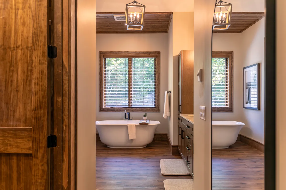 A spa-inspired ensuite bath attached to a suite, featuring a freestanding bathtub positioned beside large windows, wooden cabinetry, and an abundance of natural light. The mood is serene and luxurious, inviting relaxation and self-care rituals.