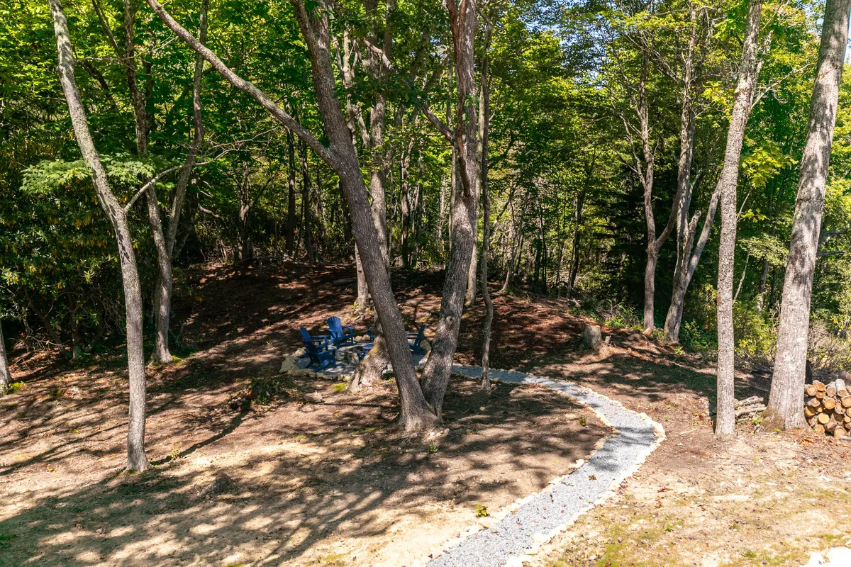 A cozy outdoor scene featuring a stone fire pit surrounded by two blue chairs, set beside a winding gravel path and tall trees. The tranquil environment is ideal for gatherings, storytelling, and enjoying s'mores under starry skies.