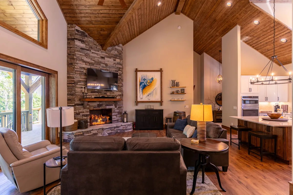 A spacious great room featuring a striking stone fireplace, comfortable seating, large windows that frame mountain views, and a visible kitchen area just beyond. The room radiates warmth and is ideal for gathering, relaxing, or entertaining guests.