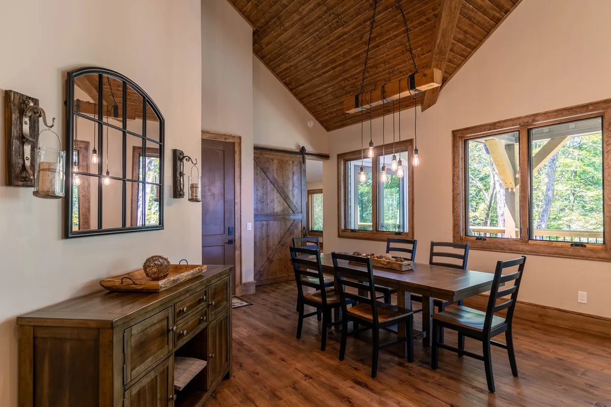 A beautiful dining area with a sturdy wooden table surrounded by chairs, flanked by large windows that offer panoramic mountain views. A decorative mirror and sideboard add touches of elegance and warmth.