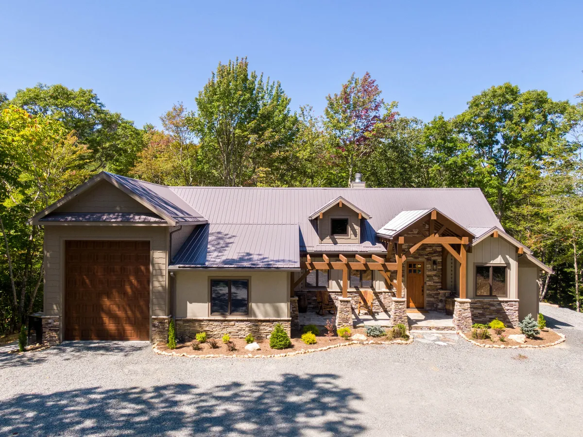 The front entrance of the cabin features sturdy wooden beams, a stone façade, a large garage door, and meticulously landscaped gardens. The driveway is visible, suggesting ample parking and easy access, ideal for visitors with electric vehicles.